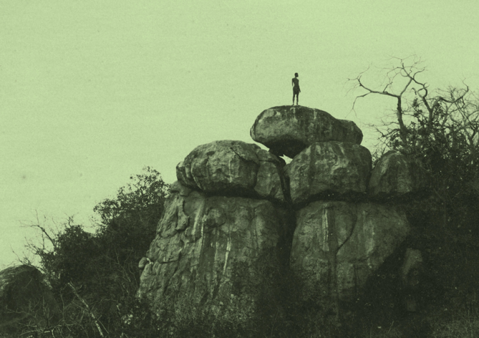 Imagem com fundo verde. Mostra um espaço de mato, com rochas e plantas secas. Um homem no topo das rochas.